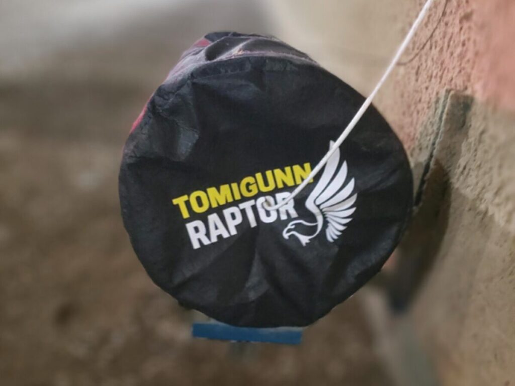 Close-up of a Tomigunn Raptor cover with a black background and a white bird logo, attached to a wall by a rope.