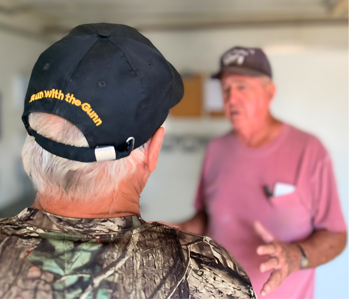 Two men are engaged in conversation indoors. The man in the foreground is wearing a black cap and a camouflage shirt, while the man in the background is in a pink shirt holding a piece of paper.