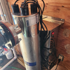 A metal UV water purifier system installed in a wooden shed, connected to pipes and an electrical outlet, with several black cables hanging from it.