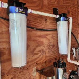 Two water filters, one larger than the other, mounted on a wooden wall. They are connected by PVC pipes, with electrical components visible beneath.