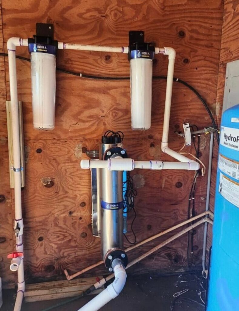 A residential water filtration system with two vertical filters, pipes, and a cylindrical stainless steel component is installed on a plywood wall. A blue water tank is visible to the right.