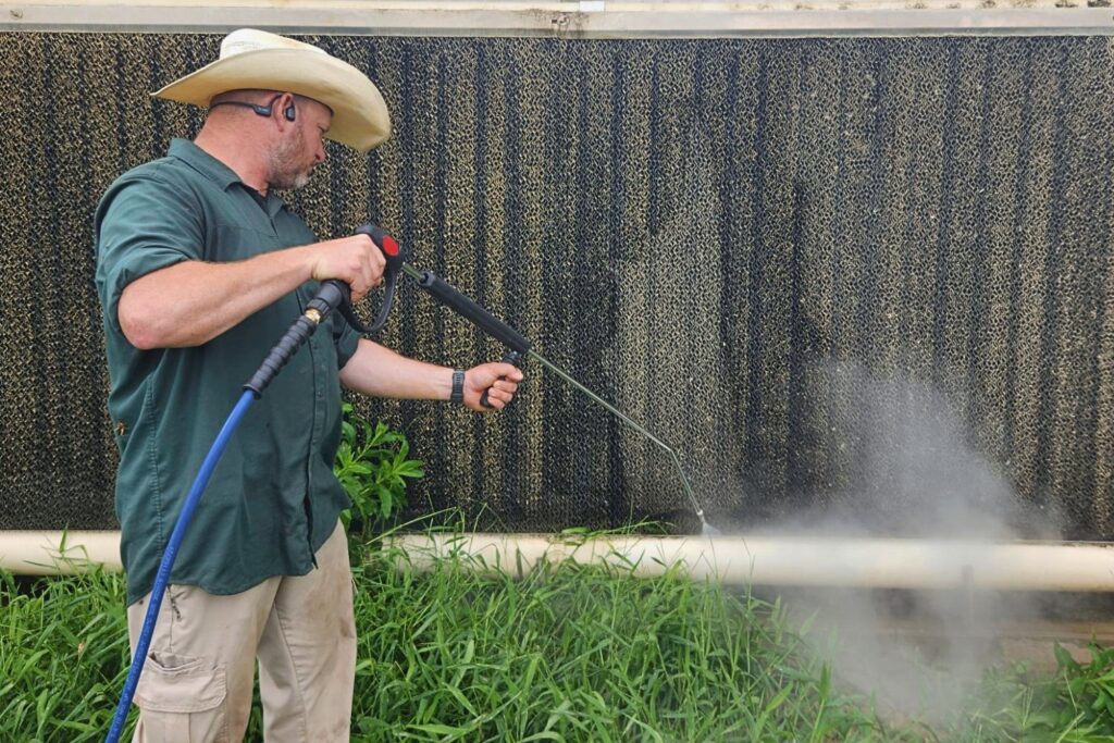 A man wearing a cowboy hat uses a pressure washer to clean a green plant area near a textured wall.